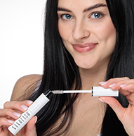 Žena elegantne drží tubu maskary Nanolash, čím predvádza svoju krásu a sebavedomie v štýlovom prostredí.