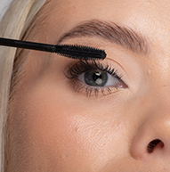 Žena si opatrne nanáša maskaru na mihalnice pomocou kefky na riasenky, čím precízne zvýrazňuje jej očný make-up.